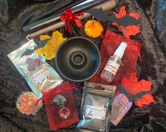 May include: A collection of Halloween-themed items, including black and gray candles tied with a red ribbon, a small orange pumpkin, and decorative leaves. Also included are a black bowl, small red pouches, and bags labeled "Samhain".