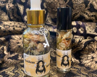 May include: Two glass bottles containing dried flowers and liquid. One has a dropper and a gold cap, the other a black cap. Both have labels with a woman's face and the words "Goddess" and "Hek". A small key charm hangs from the larger bottle.