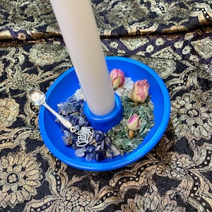 May include: A blue candle holder with a white candle, blue stones, green glass, and dried pink roses. A silver spoon and a lotus charm are also present. The holder sits on patterned fabric.