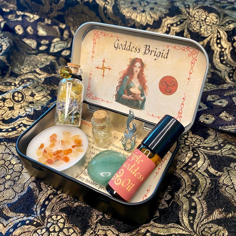 May include: A black metal tin containing various spiritual items. Inside, there's a small glass bottle with dried herbs, a candle with orange crystals, a green stone, and a roller bottle labeled "Triple Goddess Oil." The tin lid features an illustration of a woman and the text "Goddess Brigid."