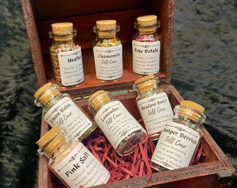May include: A wooden box filled with small glass bottles containing dried herbs and flowers. Each bottle has a cork stopper and a label with the name of the herb and the words "Self Care". The herbs include Heather, Chamomile, Rose Petals, Lemon Balm, Lavender, Meadow Sweet, and Juniper Berries.