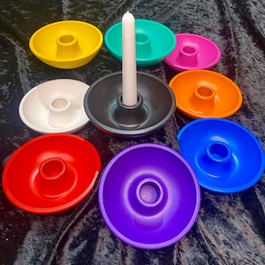 May include: A collection of colorful candle holders in yellow, teal, pink, orange, blue, red, white, black, and purple. A white candle is in the black holder. The holders are round with a central hole for the candle.