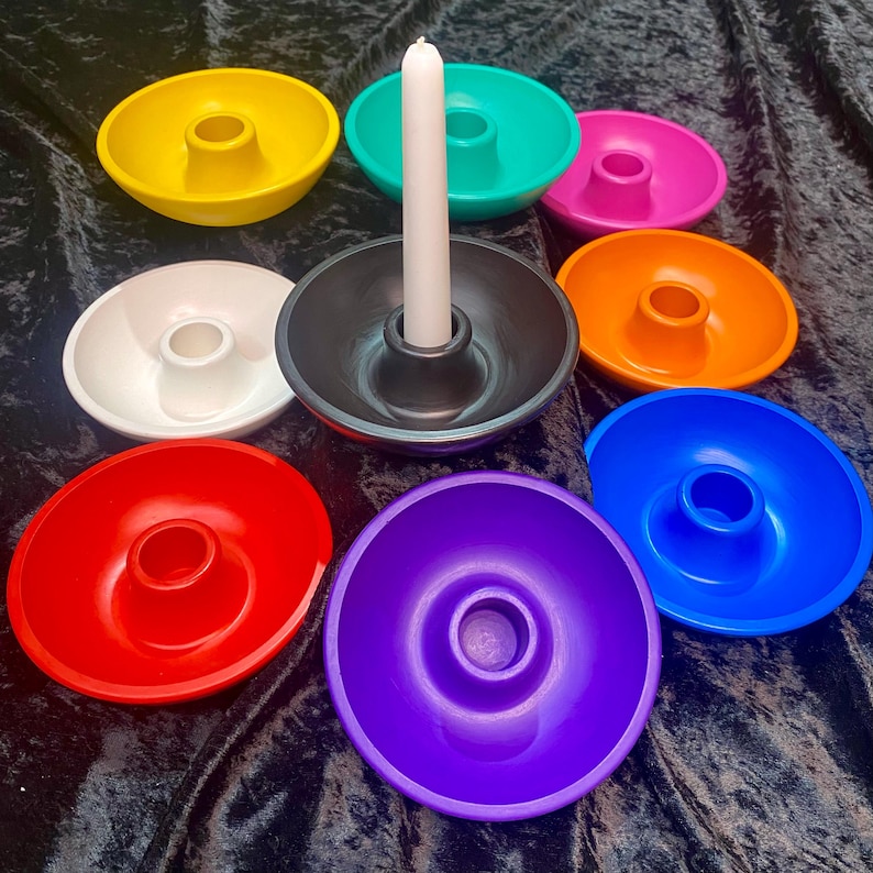 May include: A collection of colorful candle holders in yellow, teal, pink, orange, blue, red, white, black, and purple. A white candle is in the black holder. The holders are round with a central hole for the candle.
