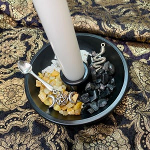 May include: A black ceramic bowl holds a white candle, small stones, and decorative charms. A silver spoon and a charm with a crescent moon and pentacle are visible. The bowl is on a gold and black patterned fabric.