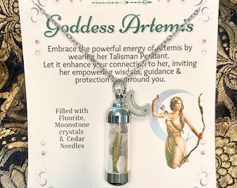 Puede incluir: Un collar plateado con un colgante de cristal transparente lleno de cristales y agujas de cedro. El colgante tiene una tapa plateada y un dije de media luna. La tarjeta detrás del collar dice "Goddess Artemis".