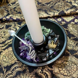 May include: A black candle holder with a white candle, filled with amethyst, green and black stones. A silver spoon and a snake charm are on the side. A gold crown charm is also present.