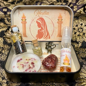 May include: A metal tin box with a Hestia theme. The box contains a small bottle of tea, a small bottle of rosemary, a candle with red and white crystals, a red stone, and a small bottle of essential oil. The box is decorated with a picture of a woman with a flame behind her.