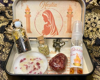Puede incluir: Una caja de metal con un tema de Hestia. La caja contiene una pequeña botella de té, una pequeña botella de romero, una vela con cristales rojos y blancos, una piedra roja y una pequeña botella de aceite esencial. La caja está decorada con una imagen de una mujer con una llama detrás de ella.