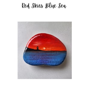 May include: A painted rock depicting a red sky, blue sea, and a silhouette of a lighthouse on a small island. The text "Red Skies Blue Sea" is written above the rock.