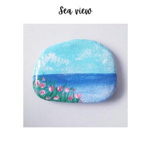 May include: A painted rock depicting a sea view. The rock is painted with a blue sky, blue water, and pink flowers. The rock is on a white background.