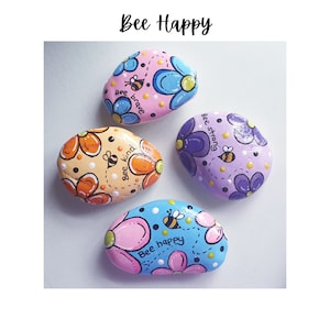 May include: Four painted rocks with bee-themed designs. Each rock has a different color background and features a bee with a message: "Bee brave", "Bee kind", "Bee happy", and "Bee strong".