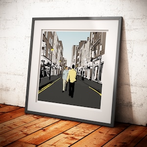 Op de afbeelding: Een ingelijste illustratie van een straat in de stad met drie mensen die over het trottoir lopen. De straat is omzoomd met gebouwen en er zijn lantaarnpalen aan de zijkanten. De mensen dragen casual kleding en lopen in dezelfde richting. De illustratie is in een zwart frame.