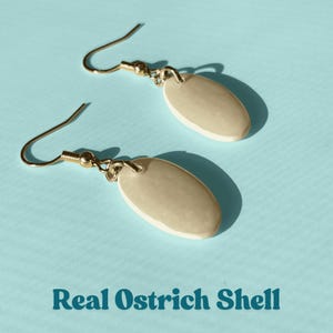 May include: A pair of earrings with gold-colored hooks and oval-shaped pendants. The pendants are a light beige color with a white border. The text "Real Ostrich Shell" is at the bottom of the image.
