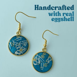 May include: Two gold-coloured earrings with a circular design. The earrings have a turquoise background with white and gold patterns, framed by a gold border. The text "Handcrafted with real eggshell" is visible in the background.