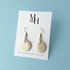 May include: A pair of cream-colored, round dangle earrings with gold-colored hooks and chain accents. The earrings are displayed on a white card with the text "MH MYROSIA" and "MyrosiaPysankyArt.Etsy.com" printed on it. The background is a light blue.