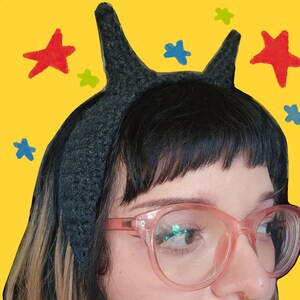 May include: A dark gray crocheted headband featuring two pointed horn-like protrusions extending from the top. The textured fabric has a dense, woven appearance, and the headband is designed to sit securely across the crown of the head.