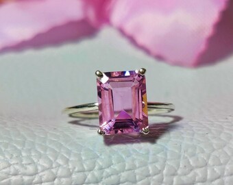 Op de afbeelding: Een zilveren ring met een grote, rechthoekige, roze edelsteen. De ring ligt op een lichtblauw, getextureerd oppervlak.