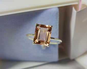 Op de afbeelding: Een ring met een grote, rechthoekige, lichtbruine edelsteen gezet in een zilveren band. De ring bevindt zich in een witte doos met een blauwe fluwelen voering.