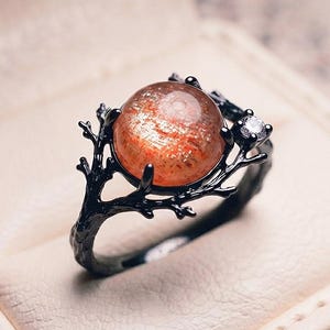 Puede incluir: Un anillo con un cabujón de piedra de sol redondo con motas naranjas, engastado en una banda de metal negro diseñada como ramas de árbol texturizadas, con una pequeña piedra preciosa transparente de talla redonda a un lado.