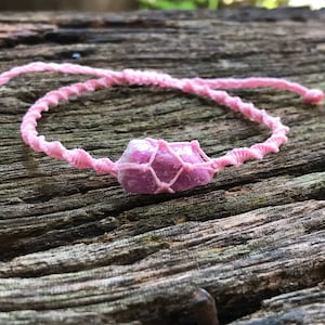 May include: Pink crystal wrapped in a pink macrame bracelet. The crystal is a light pink color and has a sparkly finish.