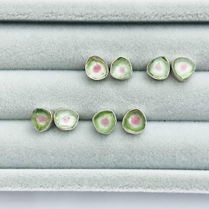 May include: Six pairs of watermelon tourmaline earrings. Each earring features a slice of green and pink tourmaline set in a silver bezel. The earrings are arranged on a gray velvet surface.