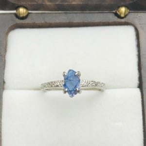 May include: A silver ring featuring a rough-cut, pale blue gemstone. The band is set with small, clear stones. The ring is presented in a wooden jewellery box with a white interior.