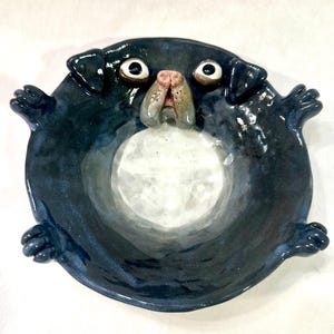 May include: A ceramic dog-shaped bowl with a dark blue exterior and a white interior. The bowl features a pug's face with large eyes, a pink nose, and small paws extending from the sides. The bowl is round and has a glossy finish.