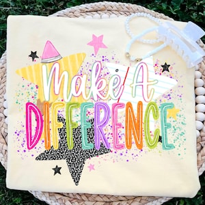 May include: A pale yellow t-shirt featuring the text "Make A Difference" in colourful, outlined letters. The design includes yellow, white, and black star shapes, pink and purple paint splatters, and a white pearl beaded accessory with a hair clip.