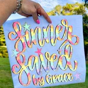Puede incluir: Camiseta azul claro con la frase "Sinner Saved by Grace" en una fuente colorida estilo tie-dye con contornos dorados brillantes. El diseño incluye estrellas de varios colores.