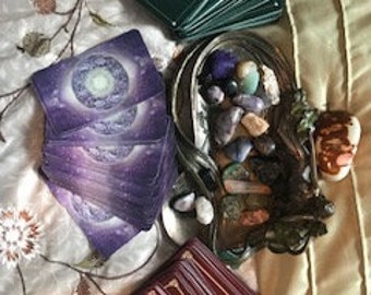 Könnte beinhalten: Drei verschiedene Tarot-Kartendecks, ein grünes Deck, ein violettes Deck und ein rotes Deck, liegen auf einem Bettüberwurf mit einem Korb voller bunter Steine.