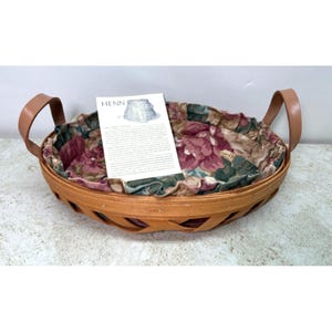 May include: Oval-shaped wooden basket with a floral fabric lining and leather handles. The basket has a light brown color and a woven design. A tag with the word "Henn" is inside the basket.
