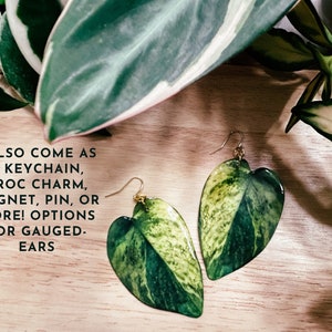 May include: A pair of green and white leaf-shaped earrings with gold hooks. The earrings are made of resin and have a realistic leaf texture. The text "I ALSO COME AS A KEYCHAIN, CROC CHARM, MAGNET, PIN, OR MORE! OPTIONS FOR GAUGED-EARS" is visible in the image.