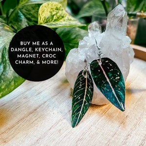 May include: Two green and white leaf-shaped earrings with a silver metal loop hanging from a white crystal rock. The text "BUY ME AS A DANGLE, KEYCHAIN, MAGNET, CROC CHARM, & MORE!" is written in black text on a black circle.