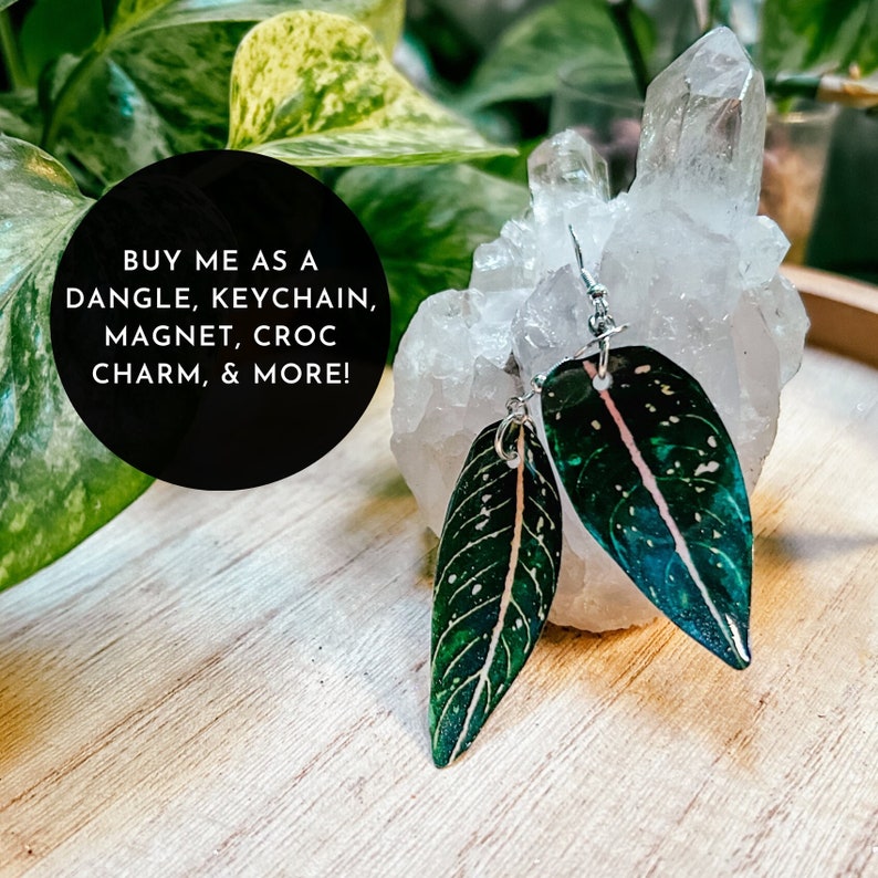 May include: Two green and white leaf-shaped earrings with a silver metal loop hanging from a white crystal rock. The text "BUY ME AS A DANGLE, KEYCHAIN, MAGNET, CROC CHARM, & MORE!" is written in black text on a black circle.