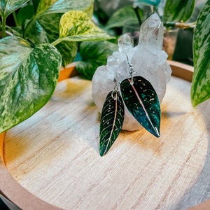 May include: A pair of dangle earrings featuring green and pink leaves with white speckles. The earrings are displayed on a white crystal and a wooden tray.