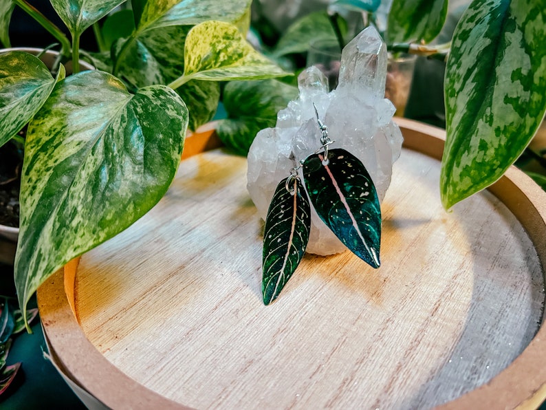 May include: A pair of dangle earrings featuring green and pink leaves with white speckles. The earrings are displayed on a white crystal and a wooden tray.