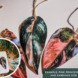 May include: Pink Princess plant leaf ornaments and earrings. The ornaments are made of polymer clay and feature a realistic pink and green leaf design. The earrings are also made of polymer clay and feature a similar design. The text "EXAMPLE: PINK PRINCESS ORNAMENT AND EARRINGS SIDE BY SIDE" is visible at the bottom of the image.