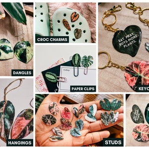May include: Assortment of handmade jewelry featuring colorful leaf designs.  The jewelry includes earrings, keychains, hanging ornaments, paper clips, pins, and shoe charms.  The text "Eat. Pray. Buys More Plants." is featured on one of the keychains.