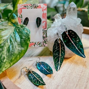 May include: Three pairs of leaf-shaped earrings with silver hooks. The earrings are made of resin and feature a green and white speckled pattern with a pink vein running through the center. The earrings are displayed on a wooden surface with a white crystal and a card with a floral pattern.