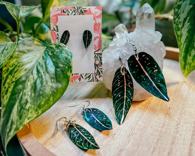 May include: Three pairs of leaf-shaped earrings with silver hooks. The earrings are made of resin and feature a green and white speckled pattern with a pink vein running through the center. The earrings are displayed on a wooden surface with a white crystal and a card with a floral pattern.
