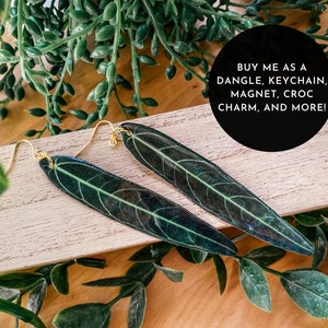 May include: A pair of leaf-shaped earrings with gold-colored hooks. The earrings are dark green with lighter green veining, resting on a light wooden surface. The image includes the text: "BUY ME AS A DANGLE, KEYCHAIN, MAGNET, CROC CHARM, AND MORE!"