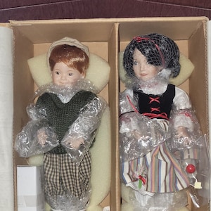 Puede incluir: Dos muñecas de porcelana en una caja. La muñeca de la izquierda es un niño que lleva un suéter verde, pantalones a cuadros y una gorra marrón. La muñeca de la derecha es una niña que lleva un vestido blanco con rayas rojas y azules, un chaleco negro y un velo blanco.