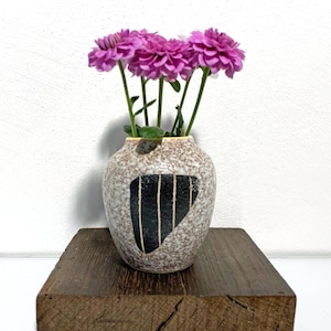 Könnte beinhalten: Eine kleine, gesprenkelte, braune und weiße Keramikvase mit einem schwarzen geometrischen Muster hält einen Strauß rosa Blumen. Die Vase steht auf einem rustikalen Holzsockel.