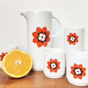 Peut inclure: Un ensemble de tasses en céramique blanche et un pichet avec un motif floral rouge et noir. Le pichet a une poignée et les tasses sont toutes de la même taille. Il y a une orange tranchée devant l'image.