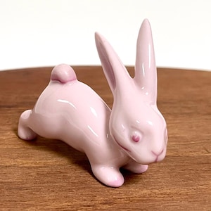 Könnte beinhalten: Kleine, rosa Keramik-Hasenfigur mit langen Ohren und einem runden Schwanz. Der Hase befindet sich in einer Ruheposition. Die Figur ist glänzend und steht auf einer Holzoberfläche.