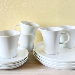 Könnte beinhalten: Ein Set aus weißen Keramikbechern und Untertassen mit passendem Milchkännchen. Die Becher und Untertassen haben ein schlichtes, elegantes Design.