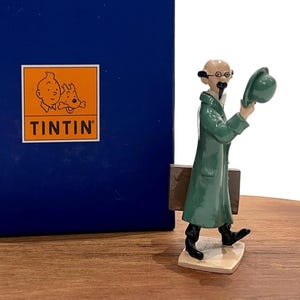 Könnte beinhalten: Figur eines Tintin-Charakters in einem grünen Mantel und schwarzen Stiefeln, der einen grünen Hut hält. Eine blaue Box mit dem Tintin-Logo befindet sich im Hintergrund. Die Figur steht auf einem beigen Sockel. Die Figur hat eine Aktentasche.