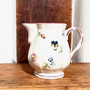 May include: A white ceramic creamer with a red rim and floral pattern. The creamer has a handle and a rounded body. The floral pattern includes red berries, blue forget-me-nots, yellow pansies, and pink flowers.