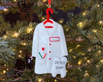 May include: A white dentist coat Christmas ornament with red accents, a red hanger, and a tooth-shaped tag. The tag reads "Dr. Hill year". The ornament includes a pocket with toothbrushes and a pen. Festive holiday decor.