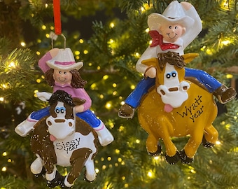 Pode incluir: Dois enfeites de Natal que retratam uma menina e um menino montando a cavalo. A menina está vestindo uma camisa rosa e um chapéu de cowboy branco com uma faixa rosa. O menino está vestindo uma camisa azul e um chapéu de cowboy branco com uma faixa vermelha. Ambos os enfeites têm o ano escrito neles. O enfeite da menina diz "Sissy year" e o do menino diz "Wyatt year".